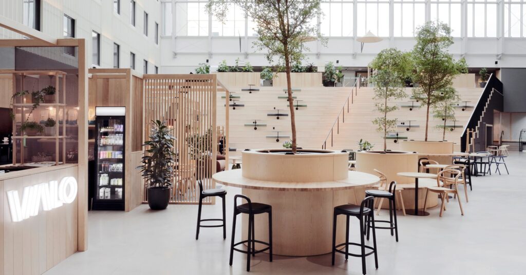 VALO Hotel & Workin valoisa atriumaula Helsingissä: puiset pöydät ja istuinportaat, puita ja lasikatto. Talossa toimii Lemkemin Helsingin toimipiste.
